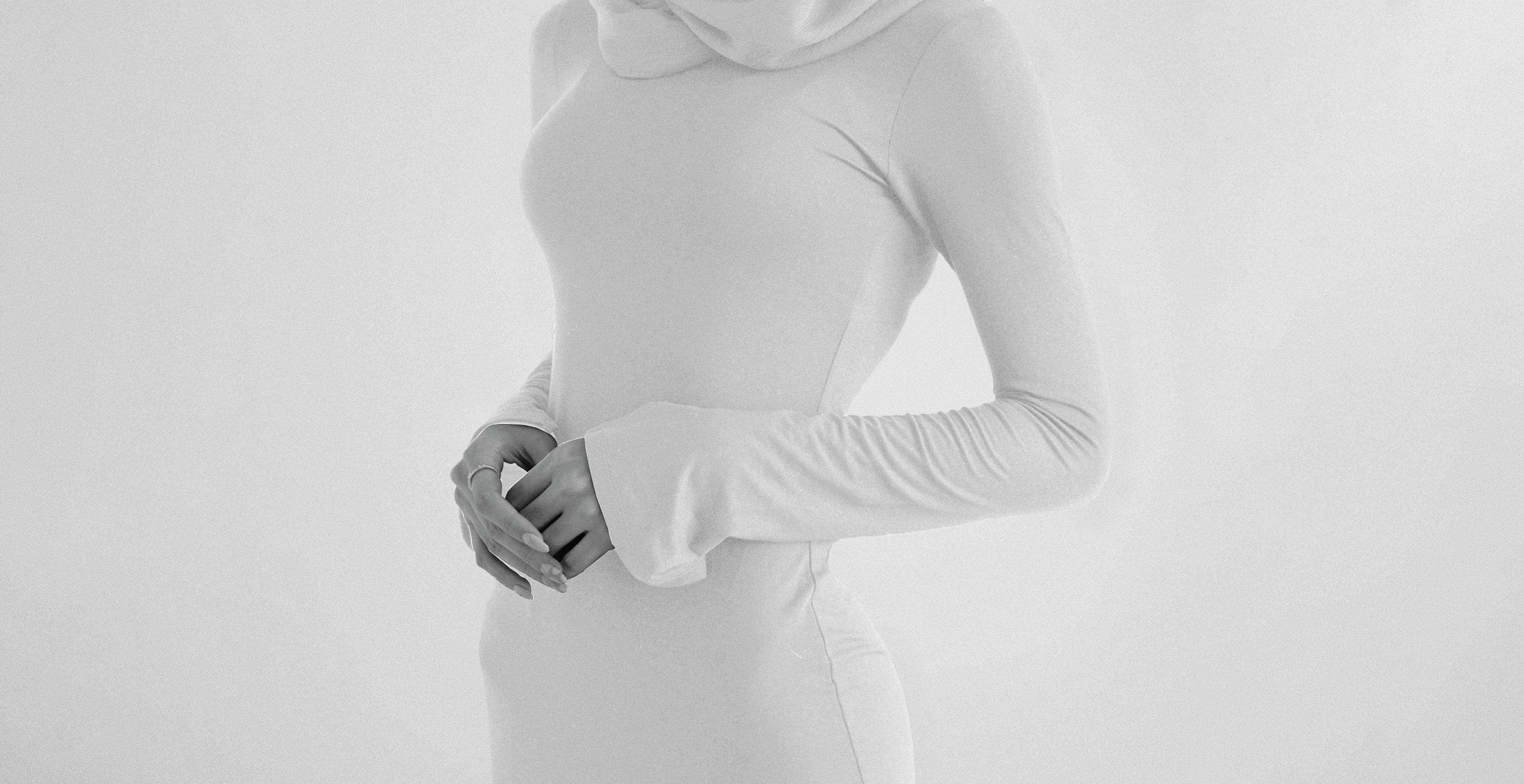 Alt ImageA black-and-white photograph showcasing Kimesca’s signature approach to luxury modest fashion. The image features a woman in a long-sleeved, high-neckline gown with a sleek, form-fitting silhouette and delicate draping. The extended sleeves and soft fabric create a refined, contemporary take on modest wear. This minimalist aesthetic reflects the essence of Kimesca—where modesty meets modern high fashion.